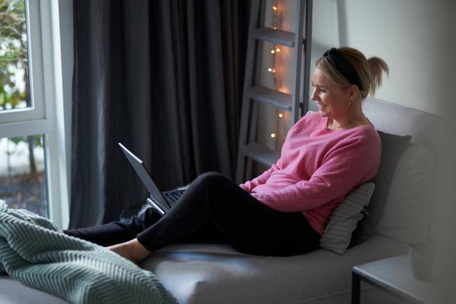 LogBuy privat - Dame i rosa genser sitter i sofaen hjemme mens hun smiler og har laptopen på fanget